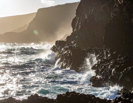 Coastal rock cliffs with waves crashing at the base, sunlit water spray, and dramatic - Powered by Adobe