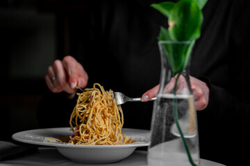 A close-up of a plate of spaghetti being eaten with a fork. A green plant in a vase decorates the...