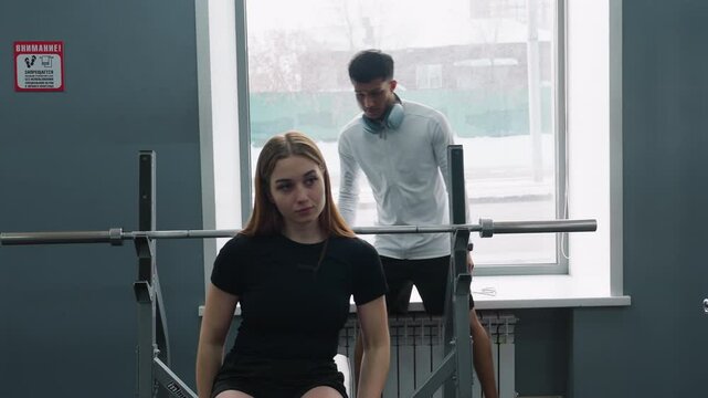 young girl lying on bench press receiving guidance from male instructor spotting barbell lift while gym environment illuminated through window with passing car outside urban fitness studio setting