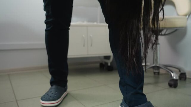 Beauty technician wearing clinical crocs moves beside spa bed as long dark hair strands fall from client onto tiled floor in bright treatment room with storage and basket under soft lighting