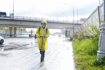 A 12 year old boy in a yellow raincoat with a backpack rides a scooter in the rain to school along...
