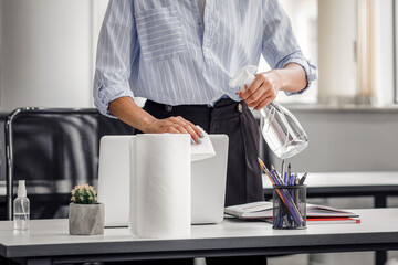 Disinfection from coronavirus in coworking place. African american young woman in protective mask working in office after covid-19 isolation, cleans laptop with antiseptic spray in interior, free