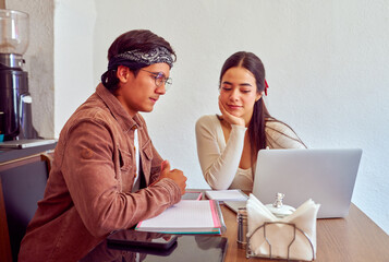 Two young students collaborating on a project in a cafe, using a laptop and notebooks