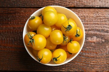 Many fresh yellow tomatoes in bowl on wooden table, top view