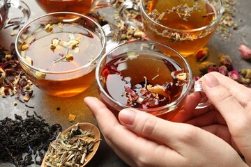 Woman with aromatic herbal tea, dry leaves and flowers at grey table, closeup