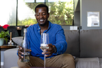 African American man sitting on gray couch in modern living room, holding wine glass and bottle