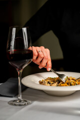 A close-up of a person dining, featuring a hand holding a fork and eating pasta from a white plate, with a glass of red wine on the table. The scene suggests a fine dining experience