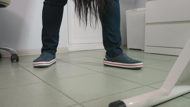 Specialist wearing clinical crocs stands beside spa bed as long dark hair strands fall from client, tiled floor, swivel chair caster and grey storage basket visible under bright overhead lighting