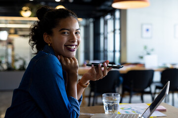 Young adult African American woman sitting at table in open office, using speakerphone near laptop