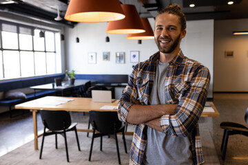 Young adult man standing with arms crossed in modern office, with laptops papers lamps, copy space
