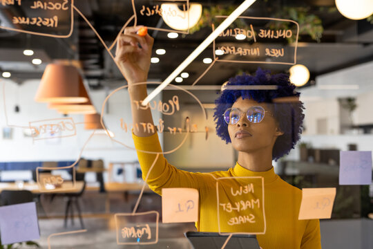 Young African American woman writing on glass board in open-plan office, with sticky notes, tablet