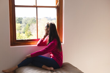 Woman sitting on window seat indoors, gazing thoughtfully out wooden-framed window, copy space