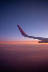 An airplane wing is captured against a stunning twilight sky filled with a gradient of blue purple orange and pink, symbolizing air travel and the serene beauty of a sunset from above the clouds.