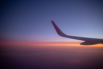 An airplane wing is captured against a stunning twilight sky filled with a gradient of blue purple orange and pink, symbolizing air travel and the serene beauty of a sunset from above the clouds.