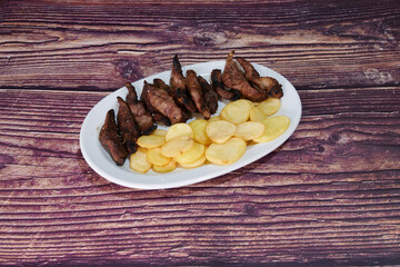 A plate of meat and potatoes on a wooden table. The meat is cut into small pieces and the potatoes are cut into wedges