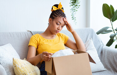 Wrong Item Concept. Sad unpset african american lady sitting on couch and unpacking cardboard box,...