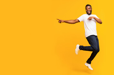 Joyful black millennial guy jumping up and pointing aside, panorama with free space, yellow studio background. Happy african american man jumping in the air, showing advertisement or text