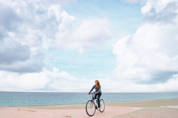Fototapeta premium Woman Riding a Bicycle on a Beachside Path with Blue Sky and Clouds..