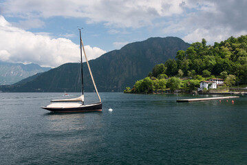 A yacht its sails down near a beautiful green shore on Lake Como in sunny weather with magnificent mountains and white clouds behind in Lenno, Lombardy, Italy