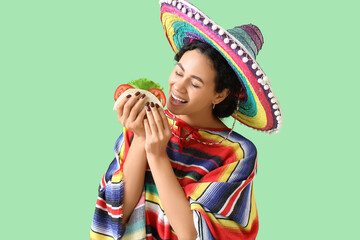 Young African-American woman in sombrero hat eating tasty taco on green background