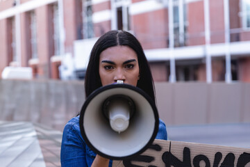 Young woman speaking into megaphone on city street, holding protest sign and wearing denim jacket
