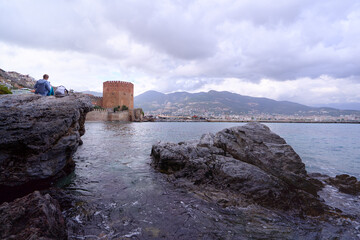 A historic brick tower stands near a rocky shore, overlooking calm turquoise waters and lush...