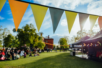 A lively outdoor festival scene with colorful bunting flags strung across a sunlit park, where...