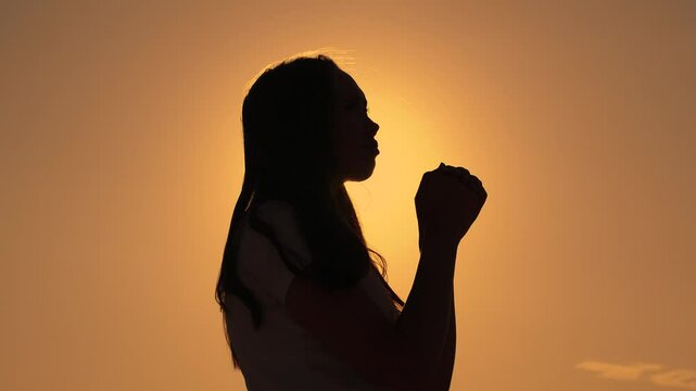 Girl on background of sky in rays of sun prayer to family and children. Relaxation and meditation in nature. Faith in god. Christian woman praying at sunset close-up. Apologize. Hope. Silhouette