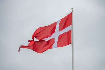 The Denmark flag flutters proudly on a flagpole, displaying its red background and white cross against a cloudy, overcast sky. The atmosphere is calm and reflective.