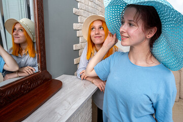 Two women are standing in front of a mirror, one wearing a blue hat