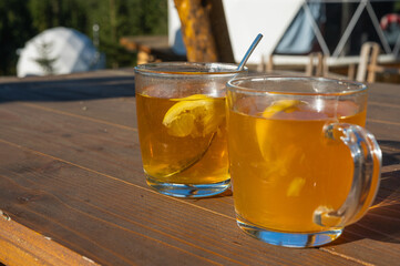 Two glass cups of hot lemon tea on a wooden table outdoors with mountains in the background. Warm sunlight, refreshing drink, natural setting.
