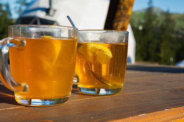 Two glass cups of hot lemon tea on a wooden table outdoors with mountains in the background. Warm sunlight, refreshing drink, natural setting.