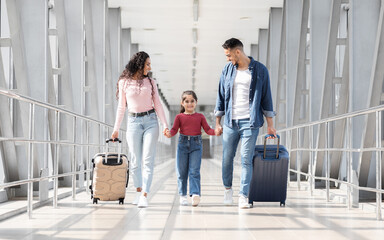 Fototapeta premium Traveling Together. Joyful Arabic Family Walking With Suitcases At Airport Terminal, Happy Middle Eastern Parents And Female Kid Carrying Luggage While Going To Flight Departure Gate, Copy Space