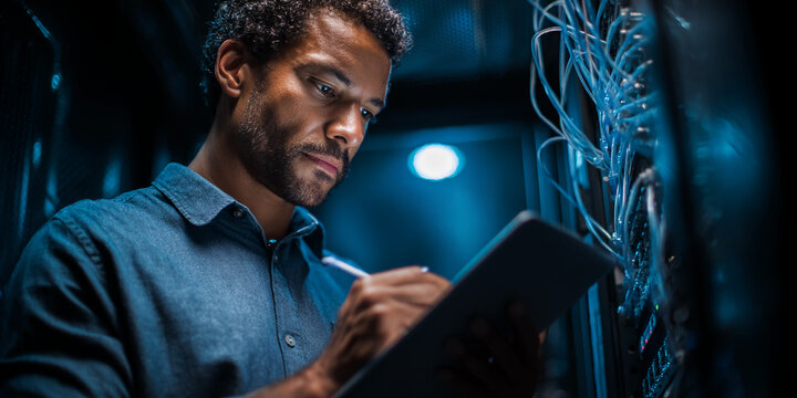 Man in blue shirt using tablet in dark server room, focused on work, depicting technological expertise and dedication