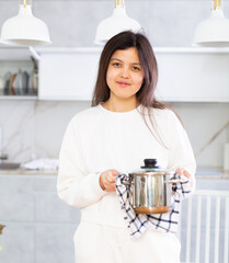 Smiling young brunette in white casual clothes standing in cozy light home kitchen, holding hot pot...