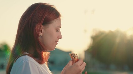 Young woman having snack outdoor. Concept fast food. Hungry girl eat burger from fast food on walk. Enjoy delicious food. Girl eat in Street. Snack in fresh food. People satisfy hunger. Teenager foods