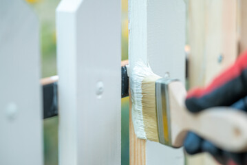 Male hand in red-black working glove painting newly made wooden fence in white colour, close up. Renovation of the garden fence by painting it using brush