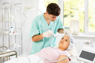 Obraz premium Young male cosmetologist performs facial moisturizing procedure with machine to young female client