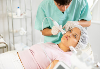 Young male cosmetologist performs hardware facial ultrasound procedure to young female patient