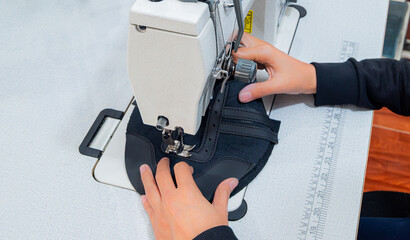 Hands sewing black synthetic leather shoe piece with industrial sewing machine in workshop, stitching footwear material with precision and control