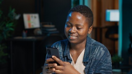Smiling woman at home texting friend on cellphone, taking break from remote job in personal office. Upbeat freelancer relaxing in apartment, talking with mate on internet using phone app, camera A
