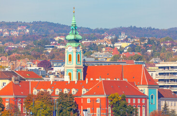 Obraz premium Vibrant view of Budapest featuring striking baroque church with turquoise clock tower and red rooftops, set against colorful hillside backdrop. Ideal for travel, architecture or Europe themed content