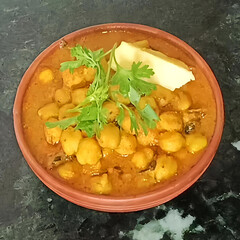 Chickpeas cooked in rich Indian curry, garnished with butter and fresh coriander, served in a clay bowl on black background.