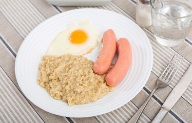 Two sausages with a fried egg and oatmeal on a white plate. Breakfast option with eggs and porridge...