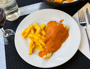 Piece of fried cod fillet covered with pureed thick escalivada sauce. Fish is garnished with French fries. Plate with hearty meal on black tablecloth.