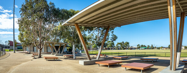 Contemporary shaded seating area with angular roof structures in an Australian public park, adjacent to an open sports field—modern civic design, urban outdoor space, and community infrastructure.