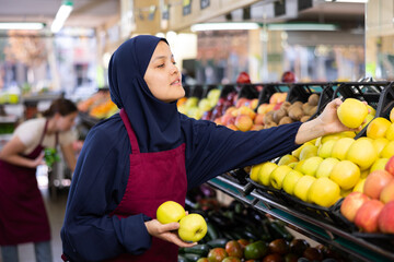 Female seller in hijab and an apron puts fresh apples on shelves in the grocery section of a supermarket