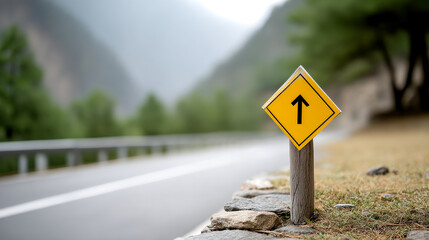 A yellow road sign with a black arrow points straight ahead along a scenic, winding road bordered by trees and mountains.