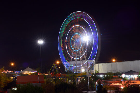 A long-exposure nighttime photo of a Ferris wheel spinning in a vibrant amusement park. The wheel forms circular light trails against the dark sky, with colorful attractions and lighting