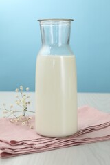 Fresh milk in jug and flowers on white wooden table against light blue background